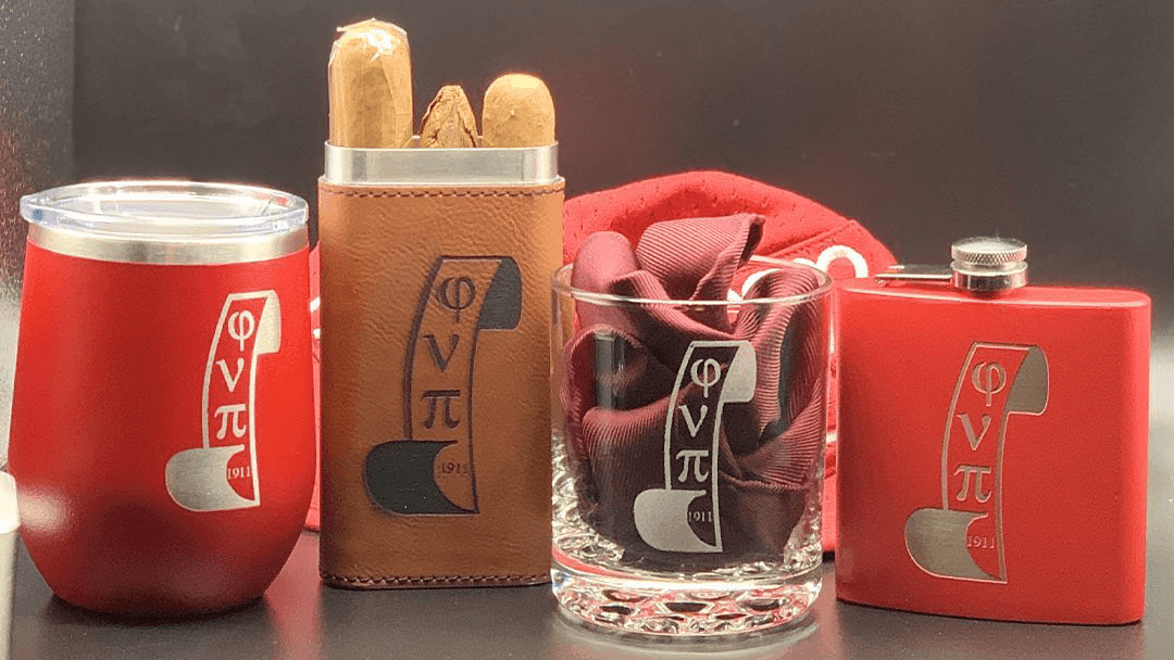 Collection of custom engraved fraternity items including a red tumbler, leather cigar case, glass, and flask,